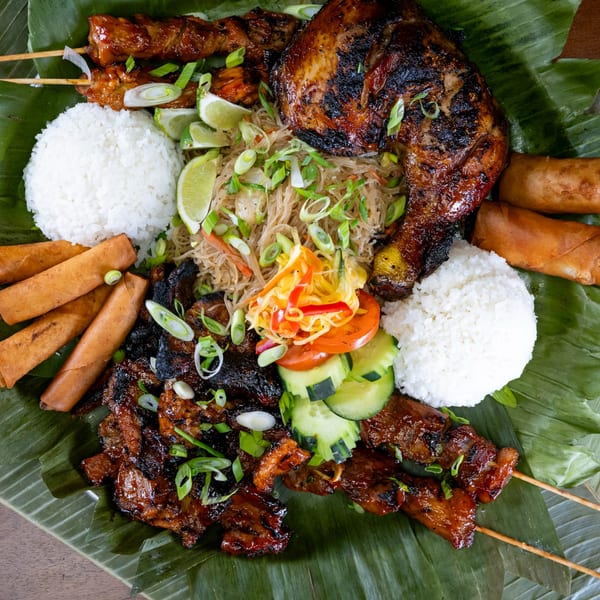 A Maemax Market Filipino feast with grilled chicken, skewers, rice, noodles, and lumpia on banana leaves.