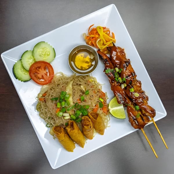 Filipino meal with chicken skewers, pancit, lumpia, pickled vegetables, and fresh cucumber and tomato.