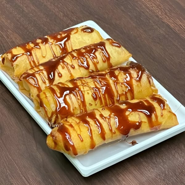 Four golden-brown Filipino turon, drizzled with sweet caramel sauce, on a white rectangular plate.