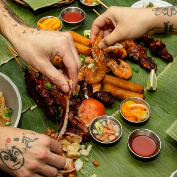 Hands sharing a Filipino boodle fight feast of grilled meats, shrimp, and lumpia on banana leaves.