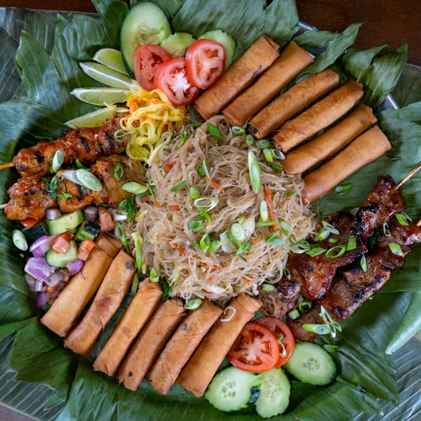 Filipino feast with pancit, lumpia, pork skewers, and fresh vegetables on banana leaves.