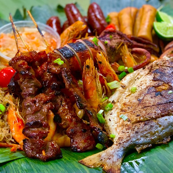 Filipino boodle fight feast with grilled fish, squid, shrimp, skewers, sausages, and lumpia on banana leaves.