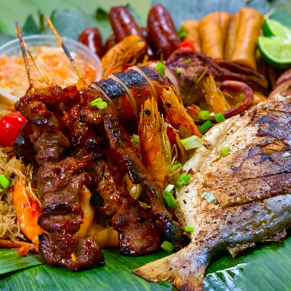 Filipino boodle fight feast with grilled fish, squid, shrimp, skewers, sausages, and lumpia on banana leaves.